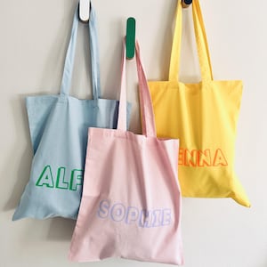 May include: Three tote bags hanging on hooks. The bag on the left is light blue with the name "ALF" in green letters. The middle bag is pink with the name "SOPHIE" in purple letters. The bag on the right is yellow with the name "ENNA" in orange letters.