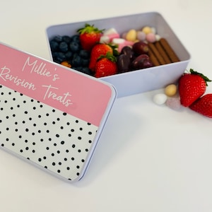 May include: A white and pink tin with black polka dots. The lid is open and the tin is filled with strawberries, grapes, chocolate, and marshmallows. The lid has the text "Millie's Revision Treats" written on it.
