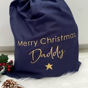 Puede incluir: Una bolsa de regalo de Navidad azul marino con letras doradas que dicen "Merry Christmas Daddy" y una estrella dorada.