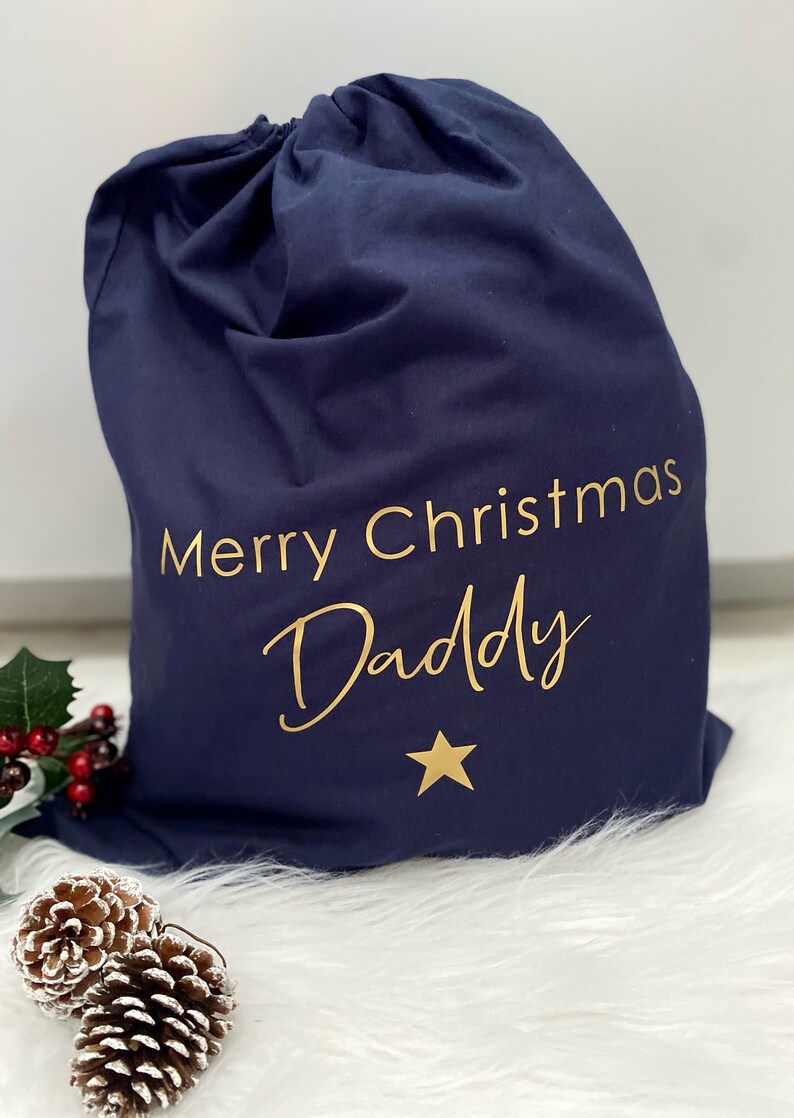 Puede incluir: Una bolsa de regalo de Navidad azul marino con letras doradas que dicen "Merry Christmas Daddy" y una estrella dorada.