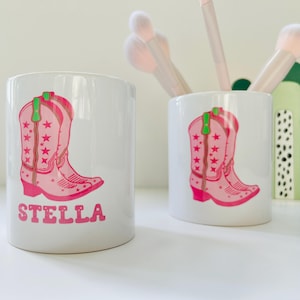May include: Two white ceramic cups with pink cowboy boots and stars printed on them. One cup has the name "STELLA" printed on it.