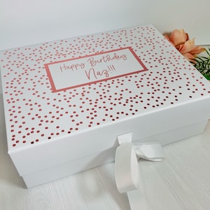 May include: A white gift box with a lid patterned with rose gold polka dots. The lid has a label with the text "Happy Birthday Naz!!!". A white ribbon is tied around the box. The box is suitable for a birthday gift.