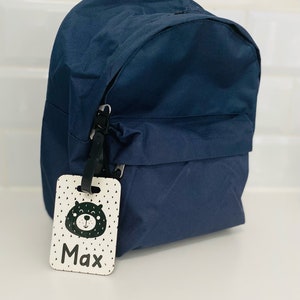 Puede incluir: Una mochila azul marino con una etiqueta de nombre blanca y negra que dice "Max" y tiene un oso de dibujos animados.