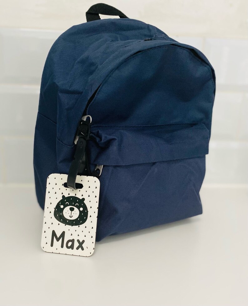 Puede incluir: Una mochila azul marino con una etiqueta de nombre blanca y negra que dice "Max" y tiene un oso de dibujos animados.