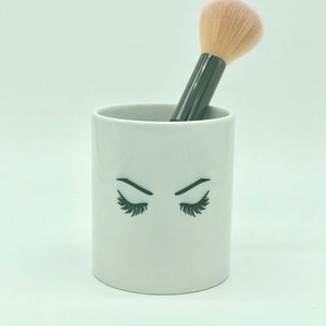 May include: White ceramic makeup brush holder with a black and white illustration of eyelashes. A black makeup brush with a pink brush head is in the holder.