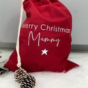 Puede incluir: Bolsa de regalo roja de Navidad con cord&oacute;n de ajuste blanco y el texto "Merry Christmas Mummy" y una estrella blanca.