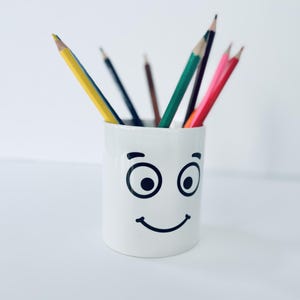 May include: A white ceramic pencil holder with a smiling face and large black eyes. The holder is filled with coloured pencils, including yellow, red, green, blue, and black.