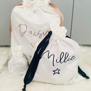 May include: Three white drawstring bags with black drawstrings. The largest bag has the name "Daisy" written in pink script with a heart. The middle bag has the name "Millie" written in black script with a star. The smallest bag has the name "Maddie" written in grey script with a heart.