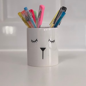 May include: White ceramic pencil holder with a cartoon face, featuring closed eyes and a small nose. The holder is filled with colourful pens, including yellow, orange, pink, blue, and green.