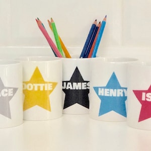 May include: Five white ceramic pencil holders with star designs and names "Grace", "Dottie", "James", "Henry", and "Isla" printed on them. The holders are arranged in a row on a white surface with coloured pencils in the centre holder.