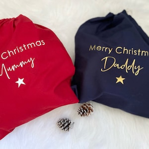 Puede incluir: Dos bolsas de regalo de Navidad, una roja y otra azul marino. La bolsa roja dice "Feliz Navidad Mam&aacute;" y la bolsa azul marino dice "Feliz Navidad Pap&aacute;". Ambas bolsas tienen una estrella dorada.