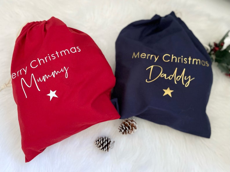 Puede incluir: Dos bolsas de regalo de Navidad, una roja y otra azul marino. La bolsa roja dice "Feliz Navidad Mam&aacute;" y la bolsa azul marino dice "Feliz Navidad Pap&aacute;". Ambas bolsas tienen una estrella dorada.