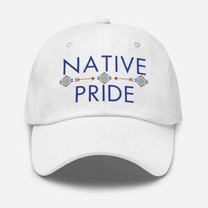 May include: A white baseball cap with the words "NATIVE PRIDE" in blue, with decorative arrows and geometric patterns in teal, red, and orange. The cap has a curved brim and a button on top.