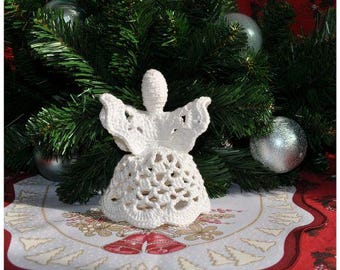 Peut inclure: Ornement d'ange en crochet blanc avec une jupe en forme de cloche et des ailes. L'ange est assis sur un chemin de table blanc et rouge avec un motif d'arbre de Noël.