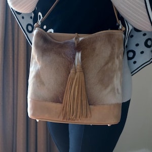 May include: A tan and brown leather shoulder bag with a fur-like texture. The bag features a light brown leather base and a matching tassel. The bag has a long strap.