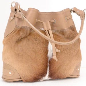 May include: A tan and brown leather bucket bag with a drawstring closure. The bag features a combination of smooth leather and textured fur, with a long shoulder strap and silver-tone hardware. The fur has a gradient effect, transitioning from brown to white.