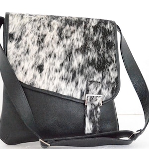 May include: Black and white cowhide crossbody bag with a black leather base and strap. The bag features a flap closure with a silver-tone buckle. The bag is designed with a long adjustable strap.