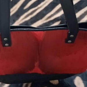 May include: A red tote bag with black handles and a black base. The bag has a rectangular shape with a rounded top and is secured with rivets. The bag is made of a soft, textured material.