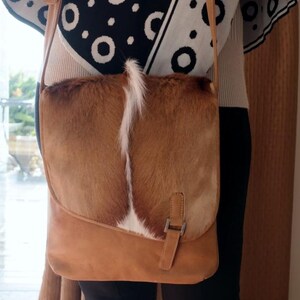 May include: A brown leather crossbody bag with a flap closure. The bag features a textured, light brown and white fur panel on the front. The bag has a long, adjustable strap and a metal buckle closure.