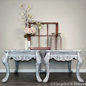 May include: Two gray painted end tables with ornate carved legs and a distressed finish. The tables are set against a white wall with a window and vases on top.