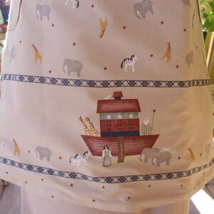 May include: Tan apron featuring a repeating pattern of elephants, giraffes, and zebras, with a border of a blue and white geometric design. The apron's lower section depicts Noah's Ark with animals and figures.