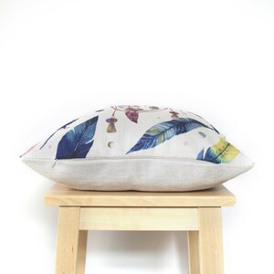 May include: A beige linen pillow with a colorful dreamcatcher and feather print. The pillow is sitting on a wooden stool.
