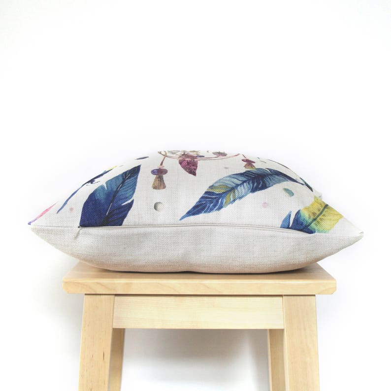 May include: A beige linen pillow with a colorful dreamcatcher and feather print. The pillow is sitting on a wooden stool.