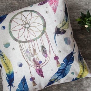 May include: A white pillow with a colorful dreamcatcher design. The dreamcatcher has feathers and beads. The background is a light beige with watercolor-like spots.