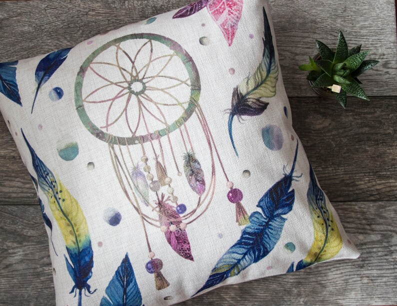 May include: A white pillow with a colorful dreamcatcher design. The dreamcatcher has feathers and beads. The background is a light beige with watercolor-like spots.