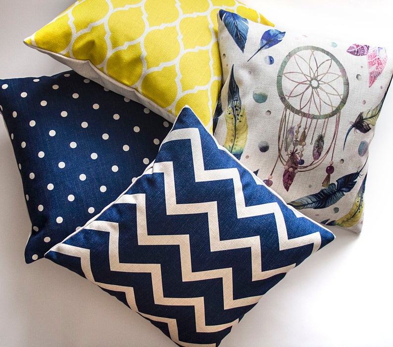 May include: Four decorative throw pillows with different patterns. One pillow has a yellow geometric pattern, one has a blue and white chevron pattern, one has a blue polka dot pattern, and one has a white background with a watercolor dreamcatcher and feathers.