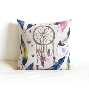 May include: A white pillow with a colorful dreamcatcher design. The dreamcatcher is surrounded by feathers in shades of blue, yellow, and purple. The pillow is perfect for adding a bohemian touch to any room.