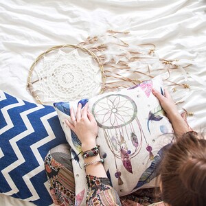 May include: A white and blue patterned throw pillow with a dreamcatcher design. The pillow is on a white bed with a white crocheted dreamcatcher hanging above it.