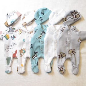 May include: Five infant footed sleepers and five matching beanies arranged in a row. The garments feature various patterns including koalas, bears, zebras, and monkeys in colors of white, light blue, and gray, each with snap button closures.