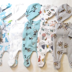 May include: Five footed sleepers and five matching beanies arranged in a row. The set includes patterns featuring blue bears, solid white, zebras, forest animals with text, and brown monkeys on gray fabric. Each sleeper has a front snap closure.