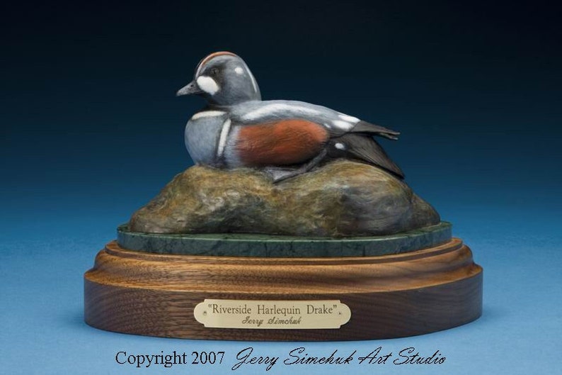 May include: A wooden sculpture of a Harlequin Duck, also known as a 'Riverside Harlequin Drake', resting on a rock. The sculpture is painted in realistic detail, with a gray, white, and orange color scheme. The sculpture is displayed on a wooden base with a green marble inlay and a brass plaque that reads "Riverside Harlequin Drake" and "Jerry Simechuk".