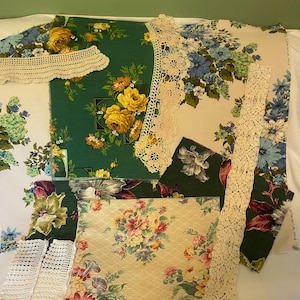 May include: A collection of assorted fabric scraps featuring floral patterns in green, blue, yellow, and cream, layered with various pieces of cream-colored crocheted lace trim. The textiles display diverse botanical prints and intricate needlework.