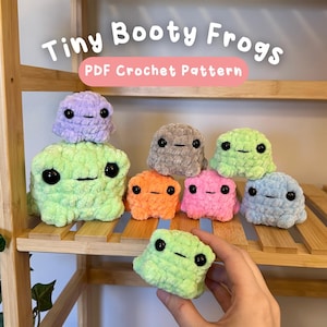 May include: A collection of small, plush, crocheted frog figures in various colors including light green, purple, gray, orange, pink, and blue. Each frog has black plastic eyes and a stitched mouth. The text Tiny Booty Frogs PDF Crochet Pattern is visible.