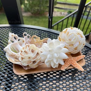May include: A wooden tray displays a collection of decorative items. Included are seashells, a white flower, a starfish, and decorative spheres. The items are in shades of white, beige, and orange, creating a coastal theme.