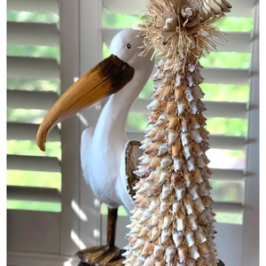 May include: A white pelican figurine stands next to a seashell Christmas tree. The tree is decorated with burlap and a white flower. The tree is on a dark brown table.