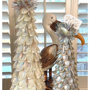 May include: Two decorative Christmas trees made of iridescent shells, adorned with silver tinsel and a tag that reads "Peace". A ceramic seagull statue is positioned behind the trees, adding a coastal theme.
