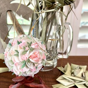May include: A white and pink floral sphere made of seashells and fabric flowers on a wooden stand. The sphere is surrounded by dried palm leaves.