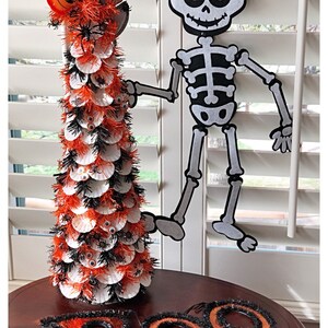 May include: A Halloween-themed tabletop tree decorated with white and orange paper cupcake liners, black and orange tinsel, and googly eyes. A felt skeleton cutout with purple eyes is hanging on a window behind the tree. The tree is on a wooden table with a black and orange tinsel "BOO" sign.