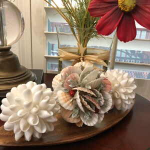 May include: Three white seashell ornaments arranged on a wooden tray. The ornaments are made of white seashells and resemble flowers. The tray is brown and has a curved shape. A vase with red flowers and green leaves is in the background.