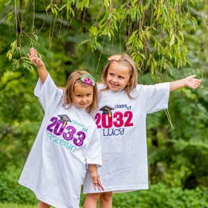 May include: Two girls wearing white t-shirts with the text "Class of 2033 Gracelyn" and "Class of 2032 Lucy" on them. The girls are standing in front of a green leafy background.