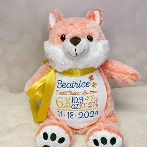 May include: A plush peach-colored fox with a white belly and black paw pads. It is wearing a yellow ribbon and a white bib with the text "Beatrice", "Freda Taylor-Lawman", "6 lb 10.9", "47 cm", "6 oz 10:27", and "11.18.2024" embroidered in blue and orange.