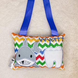 May include: A colourful tooth fairy pillow with a shark and a tooth design. The pillow has a blue ribbon for hanging and the name "Wyatt" embroidered on it.