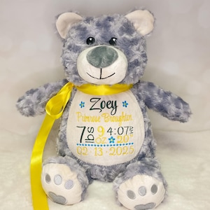 May include: A gray plush teddy bear with a yellow ribbon around its neck. The bear features a white belly patch embroidered with the name Zoey Primrose Broughton, birth details including 7 lbs 9 oz, 4:07 am, 20 inches, and the date 02-13-2025.