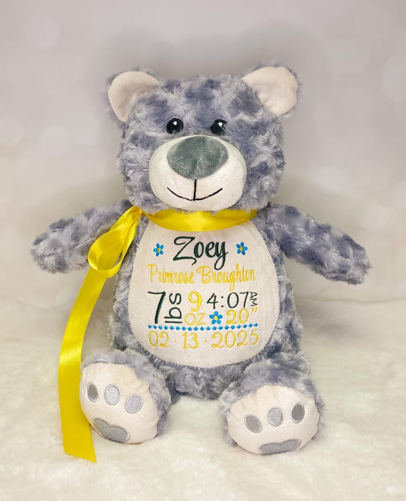 May include: A gray plush teddy bear with a yellow ribbon around its neck. The bear features a white belly patch embroidered with the name Zoey Primrose Broughton, birth details including 7 lbs 9 oz, 4:07 am, 20 inches, and the date 02-13-2025.