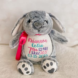 May include: A gray plush bunny with long ears and a pink ribbon. The bunny has embroidered text that reads "Rhiannon Celia" and birth details: 7 lbs 12 oz, 20.5 inches, and the date 03-15-2025.
