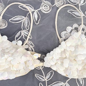 Puede incluir: Top de bikini blanco con lentejuelas y dise&ntilde;o halter. La parte superior est&aacute; cubierta de lentejuelas redondas e iridiscentes y tiene una base suave blanquecina. Las correas son finas y blancas. El top de bikini es una prenda de ba&ntilde;o.